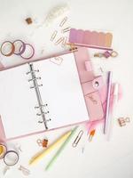 Planner with open pages on a white background and with beautiful accessories pens, buttons, pins, and colored tape. Top view of a pink planner with business stationery photo