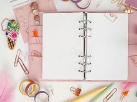 Planner with open pages on a white background and with beautiful accessories pens, buttons, pins, and colored tape. Top view of a pink planner with business stationery photo