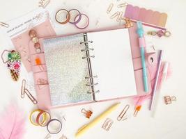 Planner with open pages on a white background and with beautiful accessories pens, buttons, pins, and colored tape. Top view of a pink planner with business stationery photo