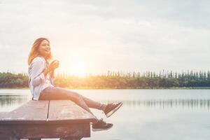 Young beautiful hipster woman holding coffee cup sitting on the lake relaxing with the fresh air and sunrise in the morning. photo