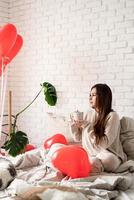 joven morena sentada en la cama celebrando el día de san valentín sosteniendo una taza de café