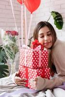 Young woman sitting in the bed celebrating valentine day embracing pile of gifts