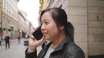 Happy Asian woman standing and having a call on street in Sweden, going out for a walk on the street in small town. Traveling abroad on long holiday. video