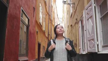 Front view of happy Asian woman walking on street in Sweden, going out for a walk on the street in small town. Traveling abroad on long holiday. Colorful building background video