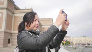 Asian woman standing and taking a picture of beautiful view of river in Sweden, standing by the river. Using smartphone taking a photo, traveling abroad on holiday. City background