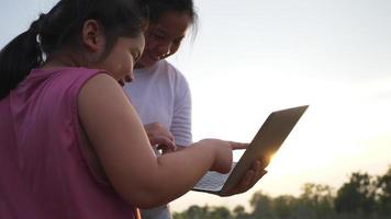 asiatisches dickes mädchen und mutter, die laptop auf dem feld benutzen, genießen sie im urlaub, stehen und spielen laptop auf gras im großen park mit vielen bäumen und sonnenlicht. schöne natur im abendkonzept video