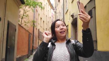 glad asiatisk kvinna som har videosamtal på smartphone på liten gata i sverige, går ut på en promenad på gatan i sverige. reser utomlands på långsemester. vacker byggnad bakgrund video