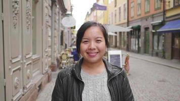 framifrån av asiatisk kvinna som står och ler på gatan i stan, går ut på en promenad på gatan i sverige. reser utomlands på långsemester. tittar på kamera koncept video