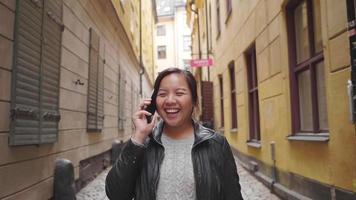 Front view of happy Asian woman walking and having a call on street in Sweden, going out for a walk on the street in small town. Traveling abroad on long holiday.
