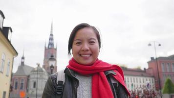 Front view of Asian woman standing and smiling on the street in town, going out for a walk to visit beautiful city in Sweden. Traveling abroad on long holiday. Looking at camera concept video