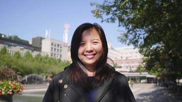 Vorderansicht einer asiatischen Frau, die in einer grünen Stadt in Schweden steht und lächelt und spazieren geht, um die Stadt in Schweden mit Bäumen und Gebäudehintergrund zu besuchen. Betrachten des Kamerakonzepts
