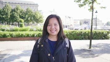 Front view of Asian woman standing and smiling in green city in Sweden, going out for a walk to visit the city in Sweden with trees and building background. Looking at camera concept video