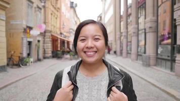 Front view of Asian woman standing and smiling on the street in town, going out for a walk on the street in Sweden. Traveling abroad on long holiday. Looking at camera concept video