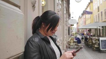 mujer asiática de pie y usando un teléfono inteligente en la ciudad, caminando por la calle en suecia. viajar al extranjero en vacaciones largas. restaurante local en el fondo de una pequeña ciudad