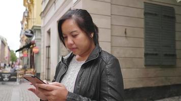 mujer asiática de pie y usando un teléfono inteligente en la ciudad, caminando por la calle en suecia. viajar al extranjero en vacaciones largas. restaurante local en el fondo de una pequeña ciudad