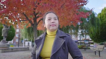Asian woman standing, looking around and smiling in the park with red and green trees. Beautiful red and green trees background, wearing winter outfits in beautiful day on the holiday, Sweden