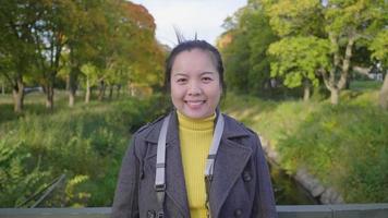 vista frontal de una feliz mujer asiática de pie y sonriendo en el puente sobre el pequeño río. un montón de árboles de fondo, vistiendo trajes de invierno mirando a la cámara en un hermoso día de vacaciones, suecia video