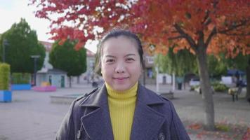 Front view of happy Asian woman standing in the park with red and green trees. Beautiful red and green trees background, wearing winter outfits looking at camera in beautiful day, Sweden video