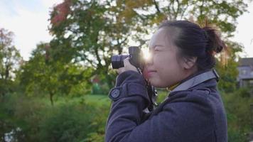 donna asiatica in piedi e scattare una foto con la fotocamera nella foresta. un sacco di alberi verdi sullo sfondo, indossando abiti invernali in una bella giornata con la luce del sole in vacanza, in svezia video