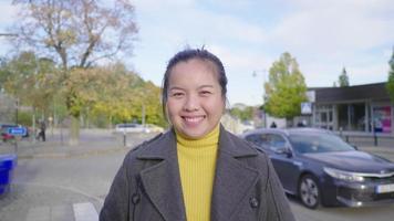Front view of happy Asian woman standing and smiling on the street. Beautiful trees background, wearing winter outfits looking at camera in beautiful day on the holiday, car moving at the back, Sweden video