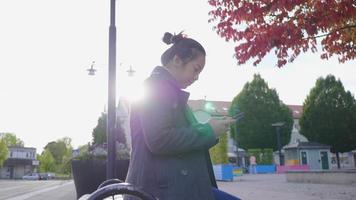 Asian woman sitting on a chair and using phone at the park with red and green trees. Beautiful red and green trees background, wearing winter outfits in beautiful day on the holiday, Sweden video