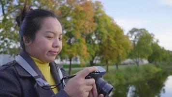 Asian woman standing and taking a picture by camera outside. A lot of green trees and long river background, wearing winter outfits in beautiful day on the holiday, Sweden video