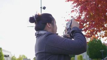 donna asiatica in piedi e scattare una foto con la fotocamera al parco. bellissimo sfondo di alberi rossi e verdi, indossando abiti invernali in una bella giornata di vacanza, in svezia video