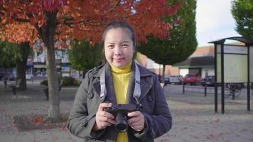 Front view of happy Asian woman standing and smiling on the street. Beautiful trees background, wearing winter outfits looking at camera in beautiful day on the holiday, car moving at the back, Sweden video