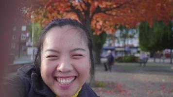 Front view of Happy Asian woman recording video herself at the park. Point the beautiful trees background. Looking at camera and taking a video selfies, wearing winter outfits in beautiful day, Sweden