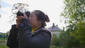 asiatisk kvinna som står och tar en bild med kameran utanför. många gröna träd och husbakgrund, bär vinterkläder i vacker dag med solljus på semestern, sverige video