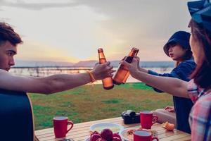 A group of Asian friends tourist drinking alcoholic beer and playing guitar together with happiness in Summer while having camping near lake at sunset photo