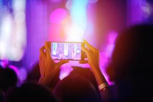 siluetas de la multitud de conciertos en la vista trasera de la multitud del festival levantando la mano y usando un teléfono inteligente para tomar fotos y videos en luces brillantes del escenario