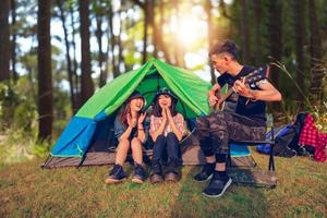 A group of Asian friends tourist drinking and playing guitar together with happiness in Summer while having camping photo