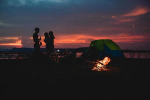 Silhouette  group of Asian friends tourist drinking and playing guitar together with happiness in Summer while having camping near lake photo