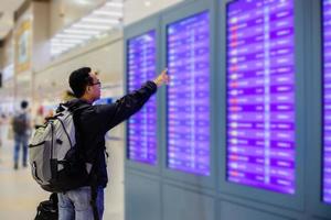 hombre asiático con mochila viajero usando el teléfono móvil inteligente para registrarse en la pantalla de información de vuelo en un aeropuerto moderno foto