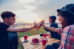 A group of Asian friends tourist drinking alcoholic beer and playing guitar together with happiness in Summer while having camping near lake at sunset photo