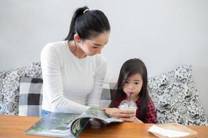 mamá está levantando un vaso de leche fría, su hija bebiendo y enseñándole a su hija a leer un libro. foto