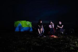 A group of Asian friends tourist drinking alcoholic beer and playing guitar together with happiness in Summer while having camping and barbecue Marshmallows on skewers over campfire near lake photo