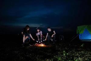 A group of Asian friends tourist drinking alcoholic beer and playing guitar together with happiness in Summer while having camping and barbecue Marshmallows on skewers over campfire near lake photo
