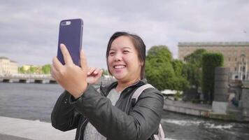 glad asiatisk kvinna har videosamtal på smartphone på liten gata vid floden i sverige, säg hej i videosamtal går ut på en promenad i sverige stad. reser utomlands på långsemester video