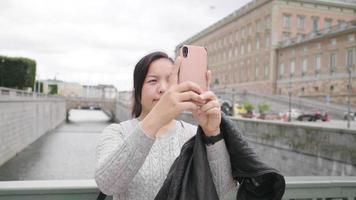 mujer asiática de pie y tomando una foto del río y la ciudad en suecia, viajando al extranjero de vacaciones. caminando por el puente y usando un teléfono inteligente para tomar una foto. hermosa ciudad en suecia video