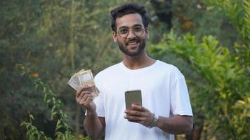 Online Shopping. Cheerful Indian Man Holding Mobile Phone And Credit Card OnOnline Shopping. Cheerful Indian Man Holding Mobile Phone And Cash, Millennial Indian Man With Cash Making