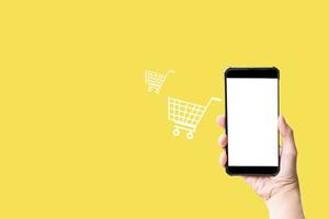 Shop online concept. Hand hold smartphone with shopping cart virtual icon on yellow background. Quarantine to save life from corona virus.
