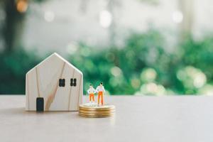 Saving money concept. Wooden home and miniature people or small model businessman figure standing on coins stack on green nature background.