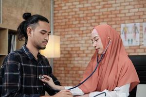 Young beautiful woman doctor is health examining male patient in office of hospital clinic and advising with a smile on medicines. This Asian medical specialist is an Islamic person wearing a hijab.