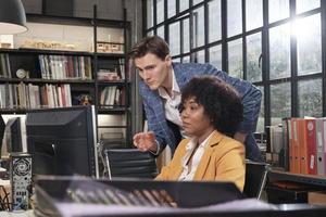 Young African American female worker sits, works with computer, brainstorming, talking, and discussing with Caucasian male colleague and partnership about business jobs in workspace office company. photo