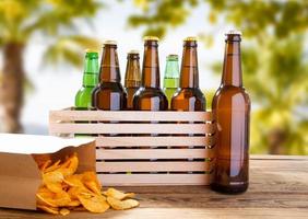 many beer bottles and pack of chips on wooden table on blurred tropical background