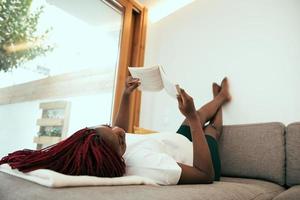 Black woman laying on the sofa reading a book with his feets on the wall