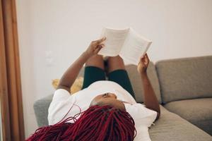 mujer negra tendida en el sofá leyendo un libro con los pies en la pared foto
