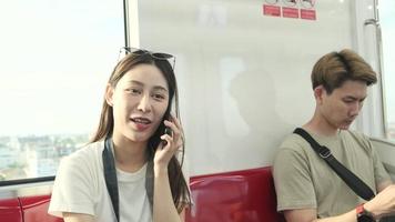 Beautiful Asian female tourist with camera sits in red seat, traveling by sky train, talking mobile phone when transporting in urban view, city passenger lifestyle by railway, happy journey vacation. video